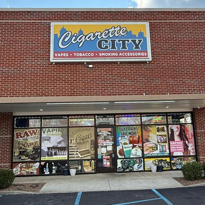 The front side view of cigarette city store (vapes, tobacco, smoking accessories) by JD stores displaying different banners of raw tobacco, vaping accessories, cigars, different types of cigarettes, and deals.