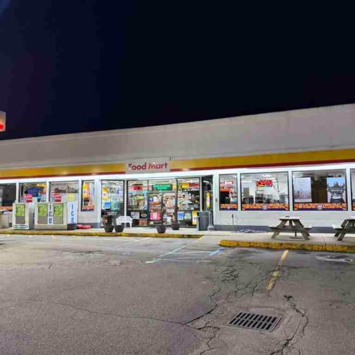 Front-side view of the Eli Shell Store by JD Stores displaying the name "Food Mart", showing edible products, a red car fueling on the left, and benches on the right for sitting.