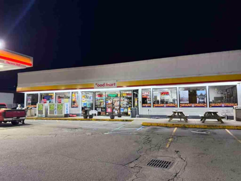Front-side view of the Eli Shell Store by JD Stores displaying the name "Food Mart", showing edible products, a red car fueling on the left, and benches on the right for sitting.